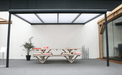 Modern outdoor dining table with benches under a metal canopy in an indoor setting.