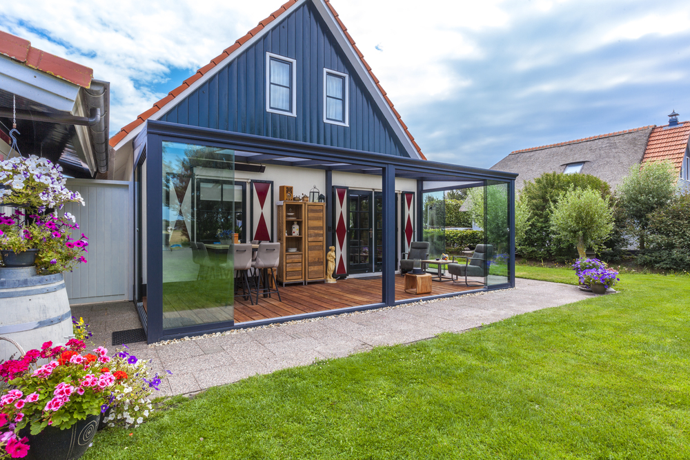 Modern extension of a house with glass walls, surrounded by greenery and flowers.