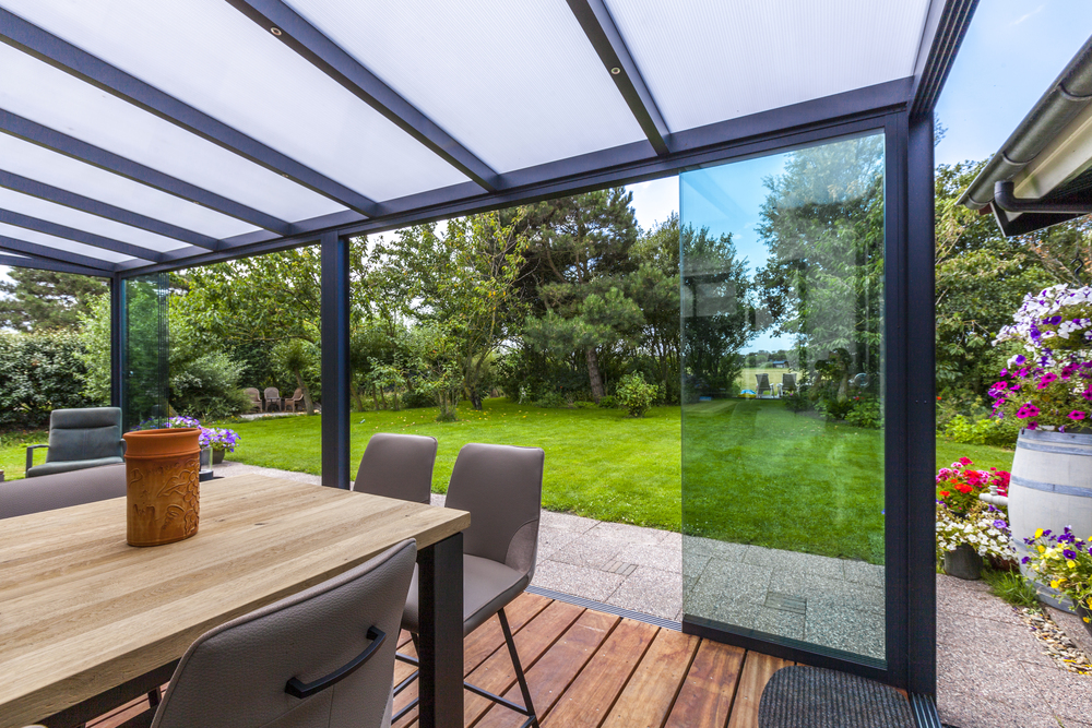 Outdoor patio with glass walls, wooden table, and chairs, overlooking a garden.