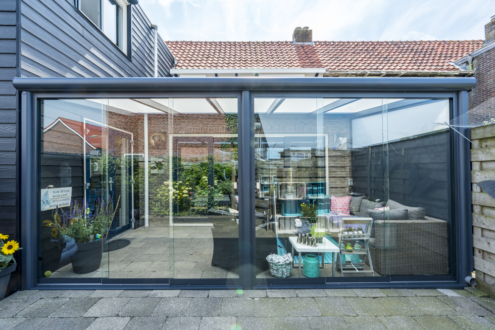 Modern glass extension connecting a house to an outdoor patio area with furniture.
