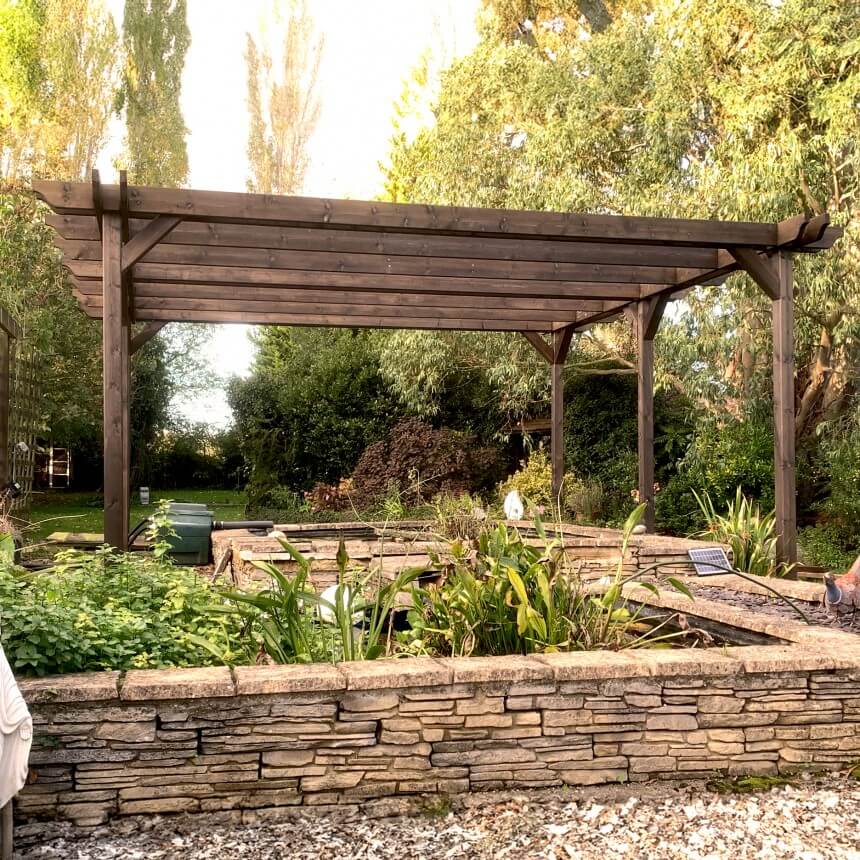 Wooden pergola over a garden with stone wall and trees in the background